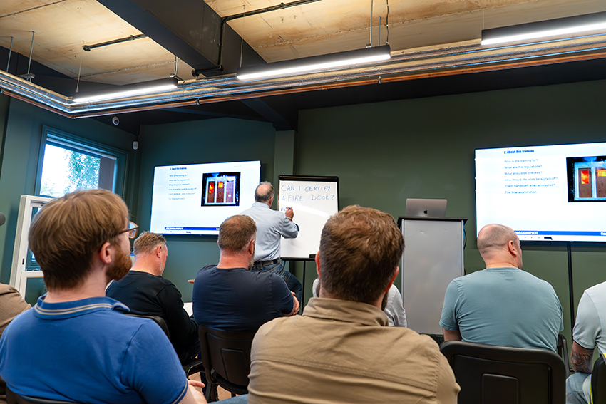Trainer delivering fire door certification course to a group of seated attendees with slides and whiteboard.